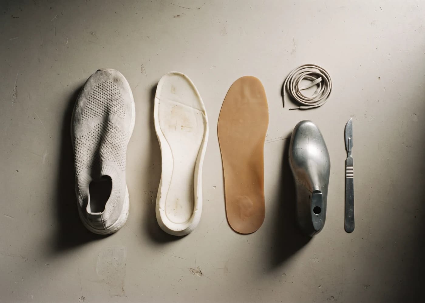 An unbranded trainer laid out in pieces on a plaster bench: knit upper, outsole, footbed, laces, last and knife